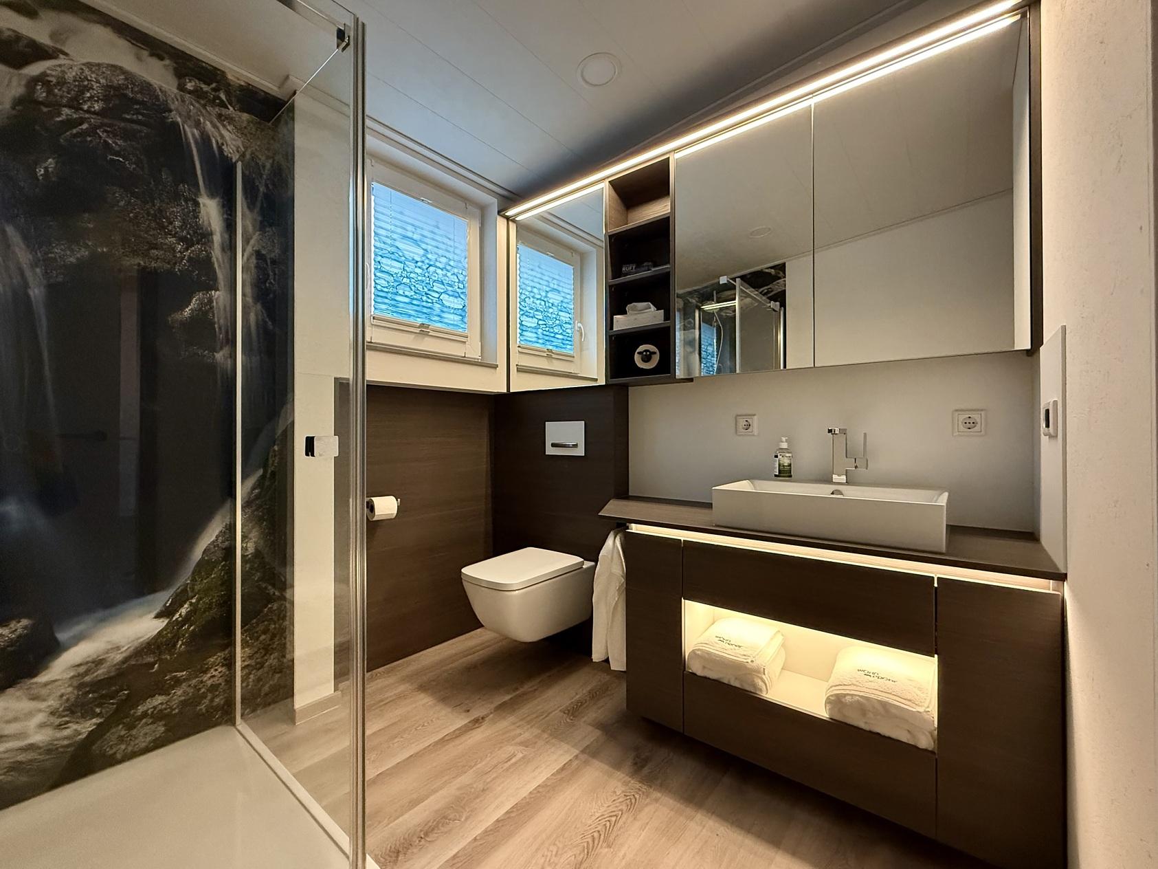 Bild zeigt Waschbeckenunterschrank mit beleuchtetem offenem Fach, Spiegelschrank &uuml;ber die gesamte Wandbreite, Wandverkleidung um das WC und Dusche mit einem Wasserfallbedruckter Duschr&uuml;ckwand fugenlos